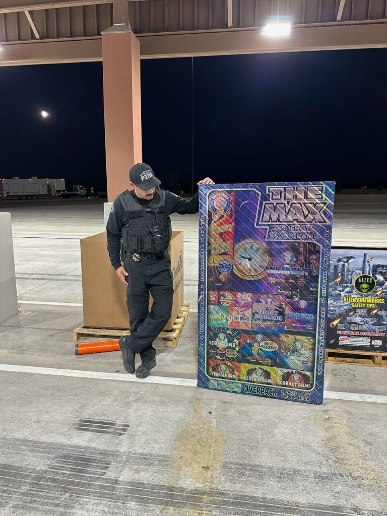 Investigator in uniform stands next to a large fireworks package labeled “The Max” during nighttime operations.