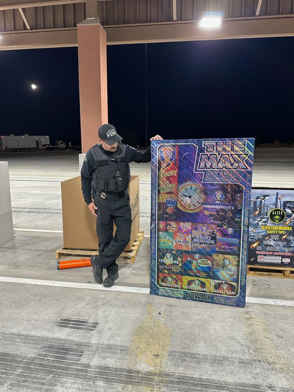 Investigator in uniform stands next to a large fireworks package labeled “The Max” during nighttime operations.