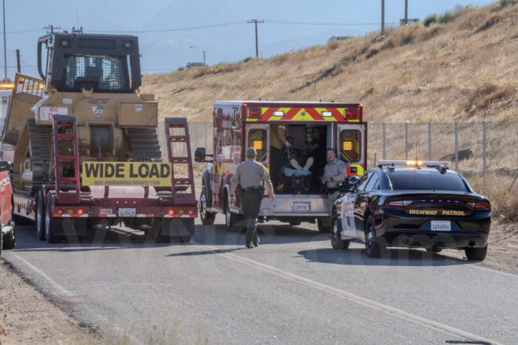 Emergency response scene on a rural road with a bulldozer on a trailer marked "Wide Load," a fire department ambulance with its back doors open and a patient inside, a sheriff’s deputy walking toward the ambulance, and a California Highway Patrol car parked nearby with lights on.