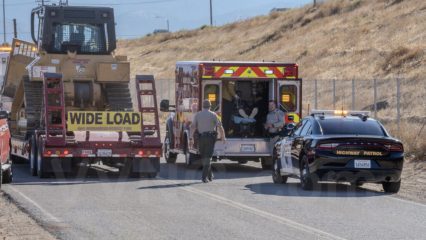 Emergency response scene on a rural road with a bulldozer on a trailer marked "Wide Load," a fire department ambulance with its back doors open and a patient inside, a sheriff’s deputy walking toward the ambulance, and a California Highway Patrol car parked nearby with lights on.