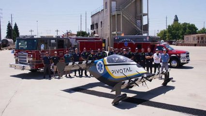 Group of firefighters and emergency responders standing in front of two fire trucks and a red pickup truck, posing with a small black, yellow, and blue single-passenger aircraft labeled “Pivotal” with a blue medical symbol on its side, in an outdoor training facility with a tall fire training tower in the background.