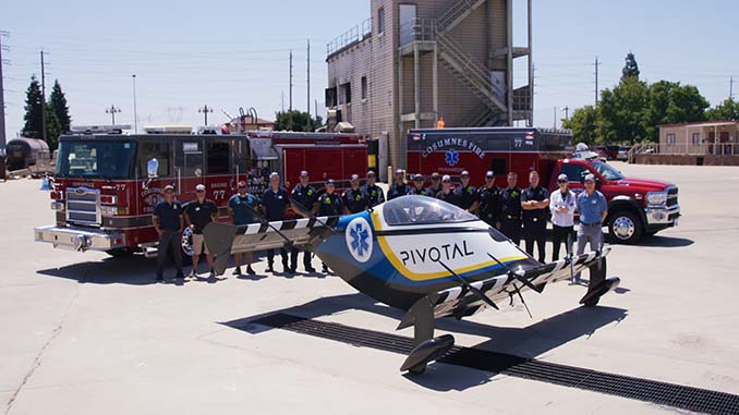Group of firefighters and emergency responders standing in front of two fire trucks and a red pickup truck, posing with a small black, yellow, and blue single-passenger aircraft labeled “Pivotal” with a blue medical symbol on its side, in an outdoor training facility with a tall fire training tower in the background.
