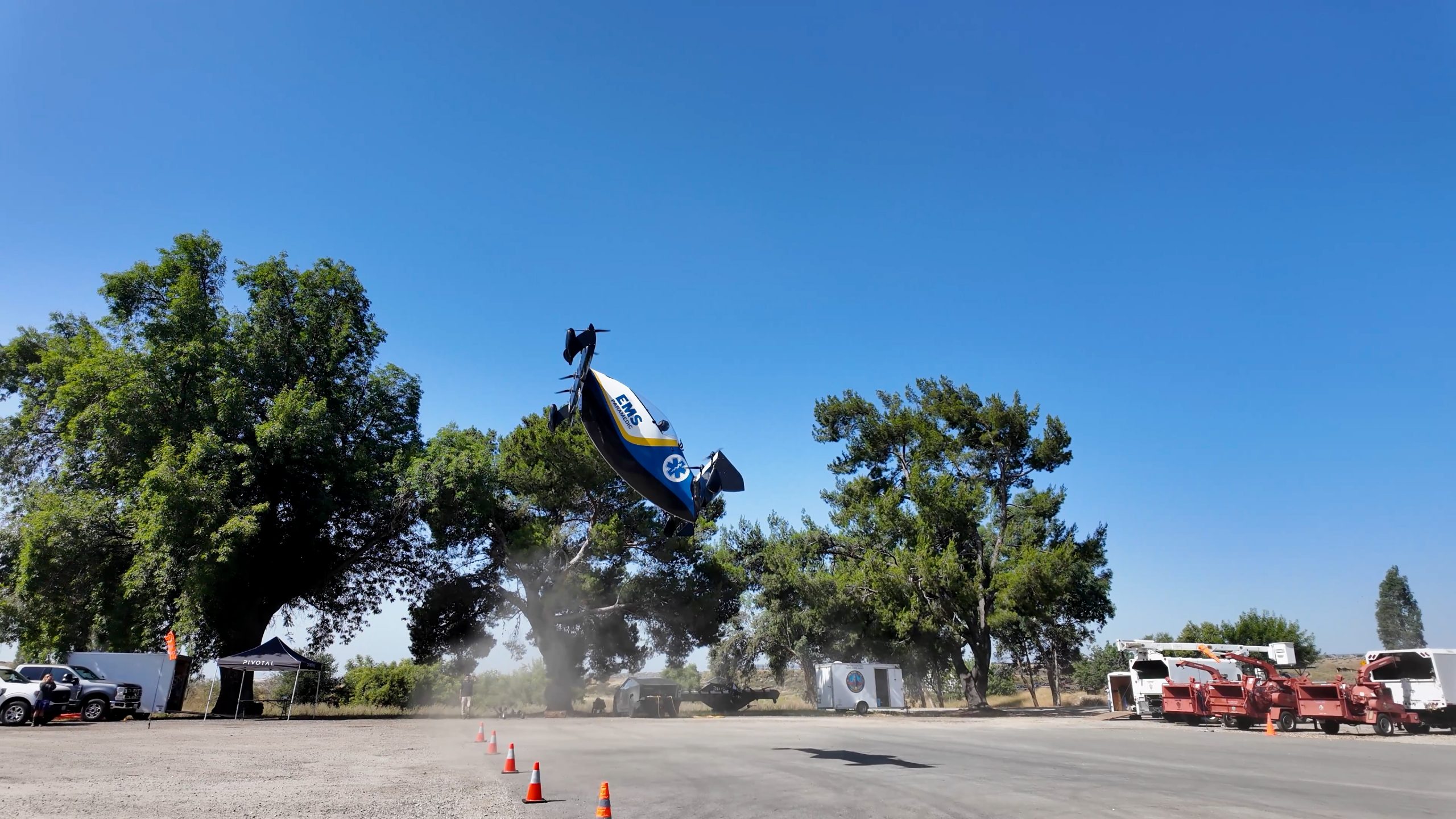 Vertical take off and landing aircraft takes off from pavement with cones on the ground and trees in the background on a clear sunny day