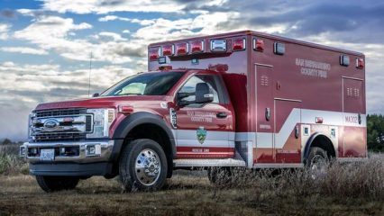 Red and white San Bernardino County Fire Department ambulance parked on grassy terrain under a cloudy sky, with emergency lights on top and department insignia on the side.