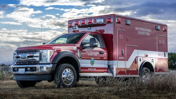 Red and white San Bernardino County Fire Department ambulance parked on grassy terrain under a cloudy sky, with emergency lights on top and department insignia on the side.