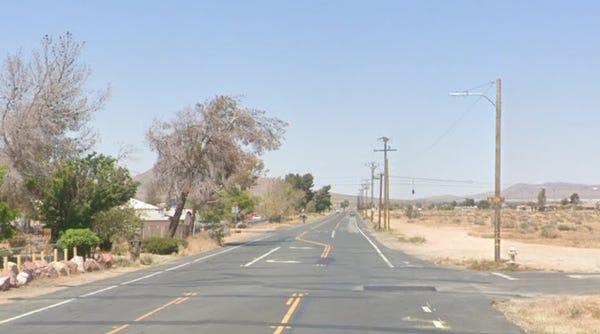 Two-lane paved road with faded yellow divider lines stretches into the distance through a rural desert community. Utility poles line the right side, with sparse houses and trees on the left and open desert landscape on the right under a clear blue sky.