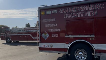 Two red San Bernardino County Fire trucks parked in a lot, with the foreground truck labeled “Hazardous Materials Response Team” and marked with a hazardous materials warning sign, and a ladder truck visible in the background under a clear sky with palm trees in the distance.