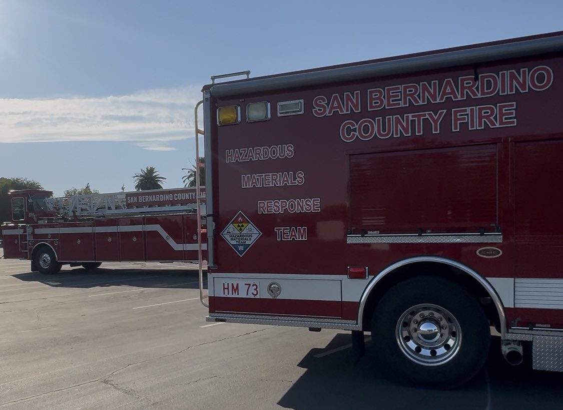 Two red San Bernardino County Fire trucks parked in a lot, with the foreground truck labeled “Hazardous Materials Response Team” and marked with a hazardous materials warning sign, and a ladder truck visible in the background under a clear sky with palm trees in the distance.