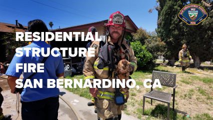A firefighter in full gear stands in front of a residential home, holding a rescued Bengal cat. Other firefighters are visible in the background. To the left, a man in blue medical scrubs faces away from the camera. Bold white text reads “RESIDENTIAL STRUCTURE FIRE SAN BERNARDINO, CA.” The San Bernardino County Fire Department logo is in the top right corner.