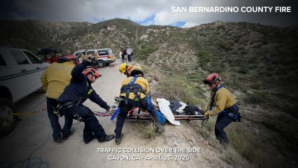 A group of San Bernardino County Fire rescue personnel, wearing helmets and safety harnesses, are pulling up a stretcher with a patient from a steep hillside during a technical rescue. The scene takes place on a dirt road overlooking a mountainous area with rugged terrain and sparse vegetation. Several emergency vehicles and bystanders are visible in the background. The image includes the text: “TRAFFIC COLLISION OVER THE SIDE – CAJON, CA – APRIL 26, 2025” and “SAN BERNARDINO COUNTY FIRE” in the top right corner.