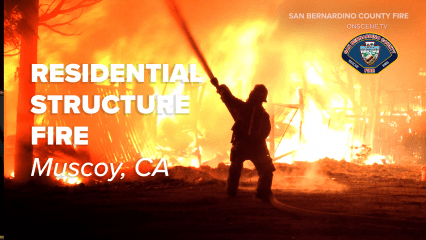 A firefighter is silhouetted against intense orange flames during a large residential structure fire in Muscoy, California. The firefighter is seen spraying water from a hose at the burning building, which is almost completely engulfed in flames. The fire illuminates nearby trees and debris, casting a bright, chaotic glow. Text on the image reads: “RESIDENTIAL STRUCTURE FIRE – Muscoy, CA.” The top right corner includes the logo of San Bernardino County Fire and the words “SAN BERNARDINO COUNTY FIRE” and “ONSCENE.TV.”