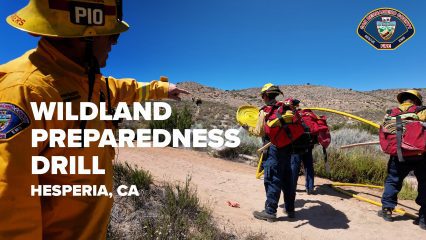 Firefighters in yellow protective gear participate in a Wildland Preparedness Drill in a desert landscape near Hesperia, California. One firefighter in the foreground points toward a trail as others carry coiled hoses and red backpacks uphill. The bright blue sky contrasts with the dry, brush-covered terrain. The San Bernardino County Fire Department logo appears in the top right corner. Bold white text on the left reads: “WILDLAND PREPAREDNESS DRILL HESPERIA, CA.”