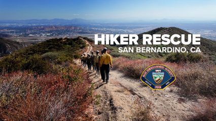 A group of firefighters in yellow and tan uniforms walk along a dirt trail on a hillside covered in brush, with a wide view of San Bernardino, California, in the background. Text on the image reads “Hiker Rescue San Bernardino, CA,” alongside the San Bernardino County Fire Department logo.