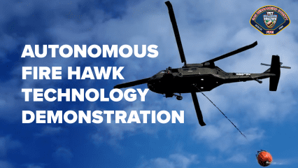 A black Fire Hawk helicopter hovers in the sky during an autonomous technology demonstration, carrying an orange water bucket suspended below by a cable. The helicopter is silhouetted against a backdrop of blue sky with scattered clouds. In the top right corner is the San Bernardino County Fire Department logo. Bold white text on the left reads: “AUTONOMOUS FIRE HAWK TECHNOLOGY DEMONSTRATION.”