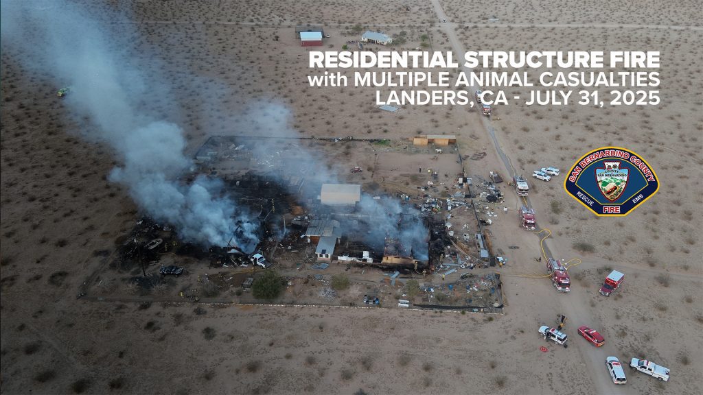 Aerial view of a large burned residential property in a desert area with thick white smoke rising from the remains, surrounded by scattered debris and multiple emergency vehicles. Text overlay reads: “Residential Structure Fire with Multiple Animal Casualties, Landers, CA – July 31, 2025” alongside the San Bernardino County Fire Department emblem.