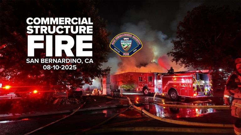 Nighttime scene of a large commercial building fire in San Bernardino, California, with flames and smoke billowing into the sky. Multiple fire engines, including one labeled “221A,” are on scene with hoses running across wet pavement. A firefighter operates equipment atop a truck while others work nearby. Text on the left reads: “Commercial Structure Fire, San Bernardino, CA, 08-10-2025” with the San Bernardino County Fire Department badge above.