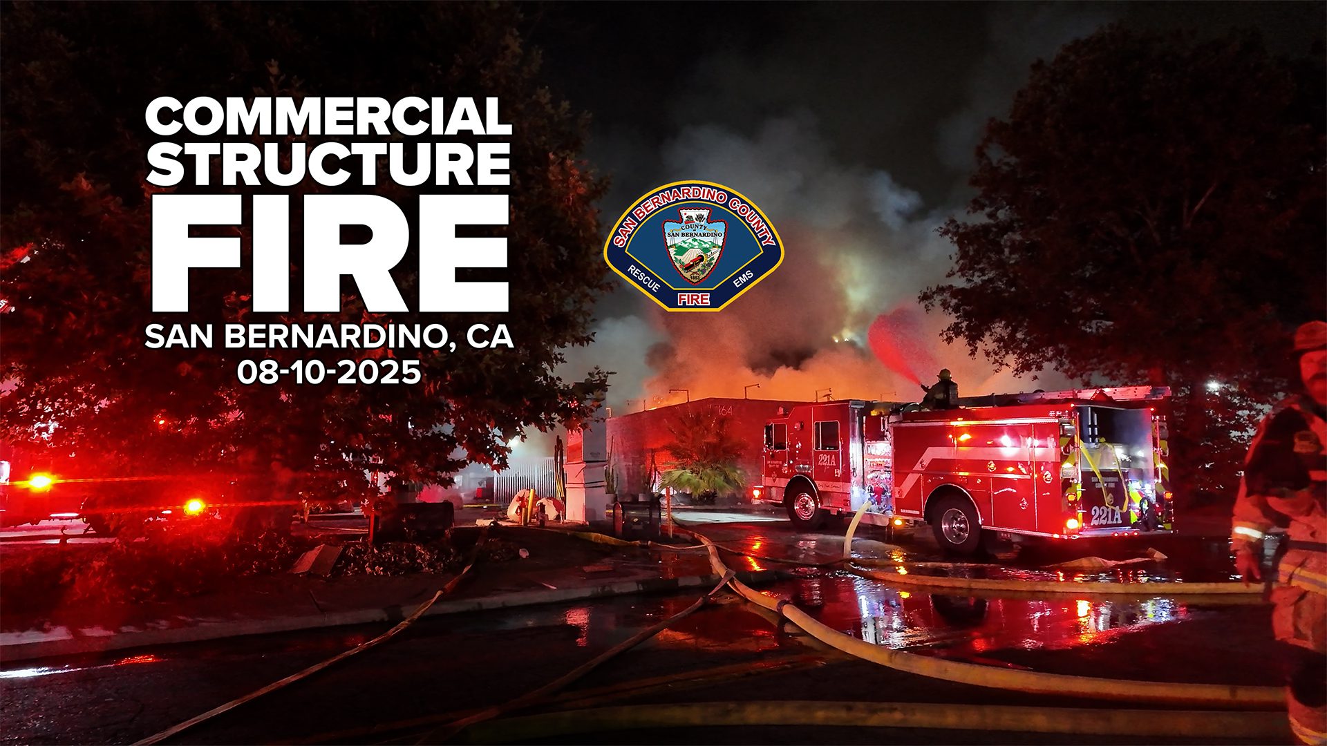 Nighttime scene of a large commercial building fire in San Bernardino, California, with flames and smoke billowing into the sky. Multiple fire engines, including one labeled “221A,” are on scene with hoses running across wet pavement. A firefighter operates equipment atop a truck while others work nearby. Text on the left reads: “Commercial Structure Fire, San Bernardino, CA, 08-10-2025” with the San Bernardino County Fire Department badge above.