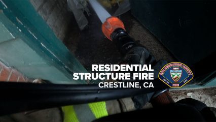 A firefighter directs a hose line spraying water inside a structure during a fire response. The firefighter’s gloved hand grips the nozzle as water streams forward. Text on the image reads, “Residential Structure Fire, Crestline, CA,” alongside the San Bernardino County Fire Department logo.