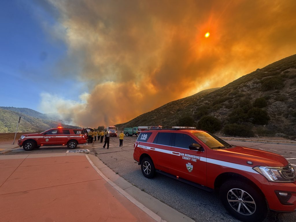 San Bernardino County Fire personnel respond to a wildfire in a mountainous area. Thick, orange smoke fills the sky as flames burn in the distance. Several red fire command vehicles with flashing lights are parked along the roadside. Firefighters in yellow protective gear stand near the vehicles, preparing for deployment. The scene is intense and urgent, set against rugged hills and an ominous, smoke-covered sun.