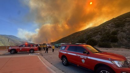 San Bernardino County Fire personnel respond to a wildfire in a mountainous area. Thick, orange smoke fills the sky as flames burn in the distance. Several red fire command vehicles with flashing lights are parked along the roadside. Firefighters in yellow protective gear stand near the vehicles, preparing for deployment. The scene is intense and urgent, set against rugged hills and an ominous, smoke-covered sun.