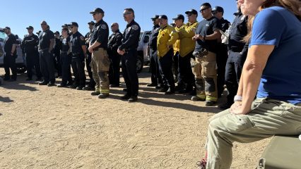 A large group of uniformed emergency personnel, including firefighters and other responders, stand in a line on a sandy surface under a clear blue sky, attentively listening to someone out of frame. Some individuals wear navy blue uniforms, while others wear yellow firefighting gear. A woman in the foreground, wearing a blue shirt, olive green pants, hiking boots, and a brown cap, sits on a green container, also listening. Several vehicles are parked in the background behind the group. The scene appears to be part of a briefing or training session held outdoors.