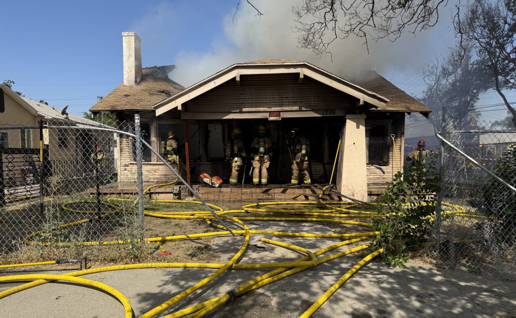 Firefighters in full gear stand on the front porch of a single-story house actively on fire, with smoke billowing from the roof and interior. The home’s address “1296” is visible above the front door. Multiple yellow fire hoses snake across the ground and through a chain-link fence in the foreground. The roof is visibly damaged, and charred sections are present around the windows and eaves. Other firefighters and emergency personnel are visible around the property, working to contain the blaze.