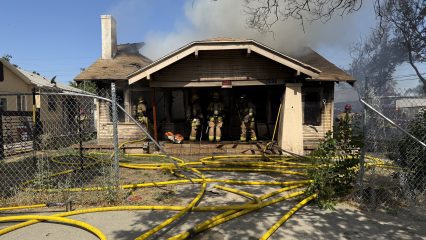 Firefighters in full gear stand on the front porch of a single-story house actively on fire, with smoke billowing from the roof and interior. The home’s address “1296” is visible above the front door. Multiple yellow fire hoses snake across the ground and through a chain-link fence in the foreground. The roof is visibly damaged, and charred sections are present around the windows and eaves. Other firefighters and emergency personnel are visible around the property, working to contain the blaze.