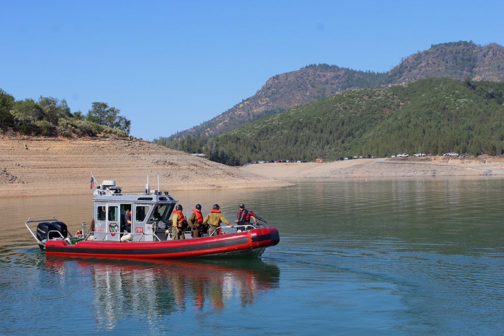 A red and gray rescue boat floats on a calm lake surrounded by mountainous terrain and pine-covered hills. Several uniformed personnel wearing life vests stand on the boat, appearing to prepare for or complete a water operation. In the background, a dirt shoreline and a line of emergency vehicles are visible along a ridge under a clear blue sky.