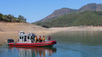 A red and gray rescue boat floats on a calm lake surrounded by mountainous terrain and pine-covered hills. Several uniformed personnel wearing life vests stand on the boat, appearing to prepare for or complete a water operation. In the background, a dirt shoreline and a line of emergency vehicles are visible along a ridge under a clear blue sky.