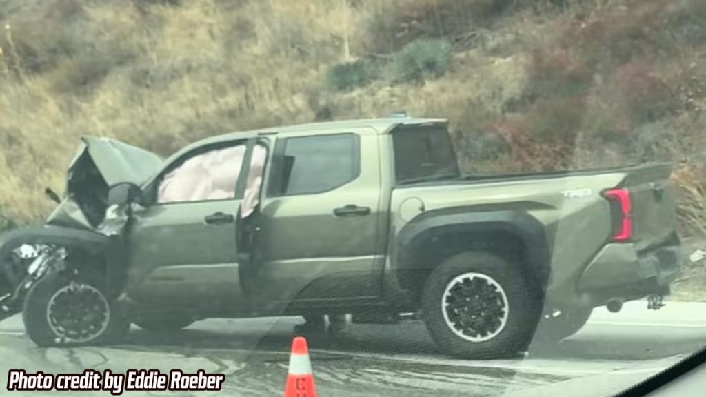 A green Toyota Tundra TRD pickup truck with severe front-end damage sits on the side of the road after a crash. The airbags inside are deployed, and an orange traffic cone is placed nearby. Dry grass covers the hillside in the background. Photo credit is given to Eddie Roeber in the bottom corner.