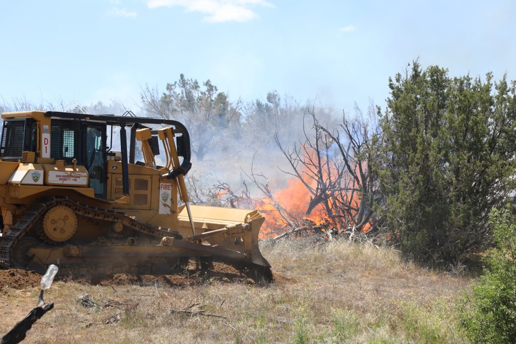 A yellow bulldozer labeled with "FIRE" and San Bernardino County Fire Department insignia creates a firebreak by clearing vegetation near an active brush fire. Flames and smoke rise from burning dead trees and shrubs in the background, while green vegetation remains nearby. The scene takes place in a dry, grassy area under a clear sky as part of wildfire containment efforts.