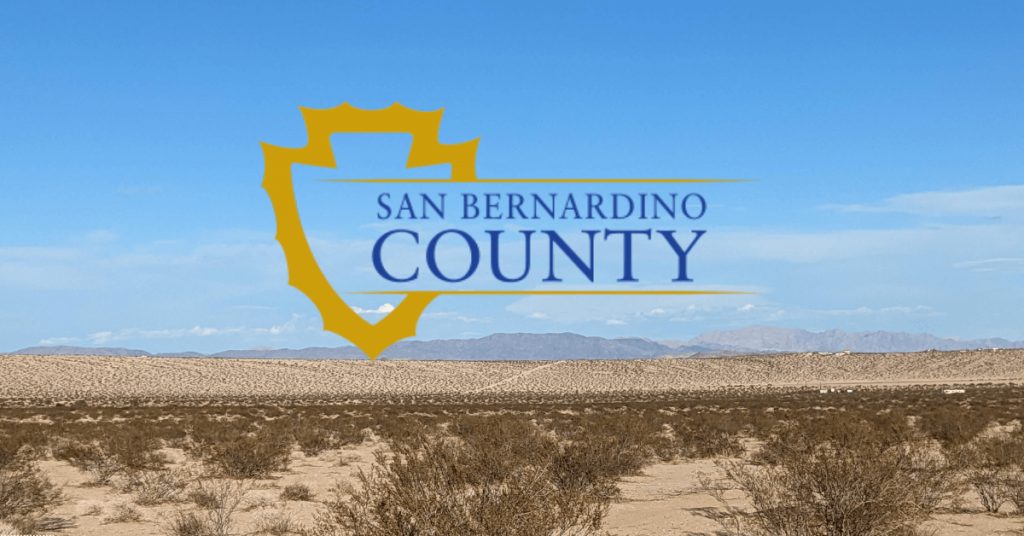A scenic desert landscape under a clear blue sky with sparse vegetation and distant mountains in the background. Superimposed in the center of the image is the official yellow and blue logo of San Bernardino County, featuring a stylized shield shape and the text "SAN BERNARDINO COUNTY" in capital letters.