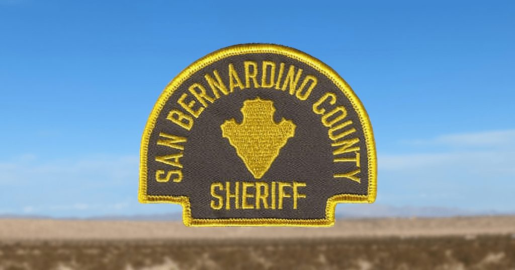 Embroidered patch for the San Bernardino County Sheriff, featuring a yellow border and text with a yellow county-shaped emblem in the center on a dark brown background, displayed against a blurred desert landscape and blue sky.