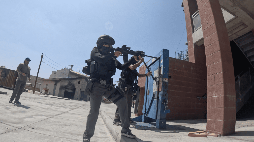 Law enforcement officers participate in an active shooter training drill outdoors under clear blue skies. One officer in tactical gear aims a rifle while another uses a battering ram to breach a heavy metal training door mounted on a red brick wall. A third individual in casual attire observes nearby. The setting includes various buildings and training structures, indicating a specialized training facility.