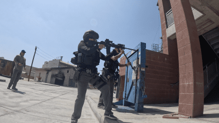 Law enforcement officers participate in an active shooter training drill outdoors under clear blue skies. One officer in tactical gear aims a rifle while another uses a battering ram to breach a heavy metal training door mounted on a red brick wall. A third individual in casual attire observes nearby. The setting includes various buildings and training structures, indicating a specialized training facility.