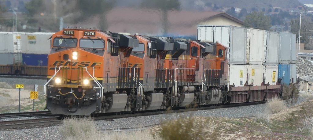 A BNSF freight train with four orange locomotives traveling on a curved track, pulling stacked cargo containers. Signs with track information are visible along the side, and dry grass and shrubs are in the foreground.