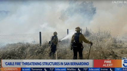 Two CAL FIRE firefighters battle a brush fire near a chain-link fence in San Bernardino during the Cable Fire. Thick smoke fills the air, partially obscuring the background. One firefighter uses a hose to spray water on the flames, while the other holds a fire tool and monitors the area. The news banner below indicates a fire alert and warns that the fire is threatening structures. Wild vegetation and dry grasses cover the ground, highlighting the fire risk.