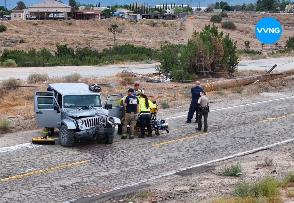 Emergency responders assist an injured person on a stretcher beside a gray Jeep Wrangler with heavy front-end damage on a cracked rural road. A downed power pole lies on the roadside, while firefighters and law enforcement officers work at the scene. The VVNG news logo is visible in the top right corner.