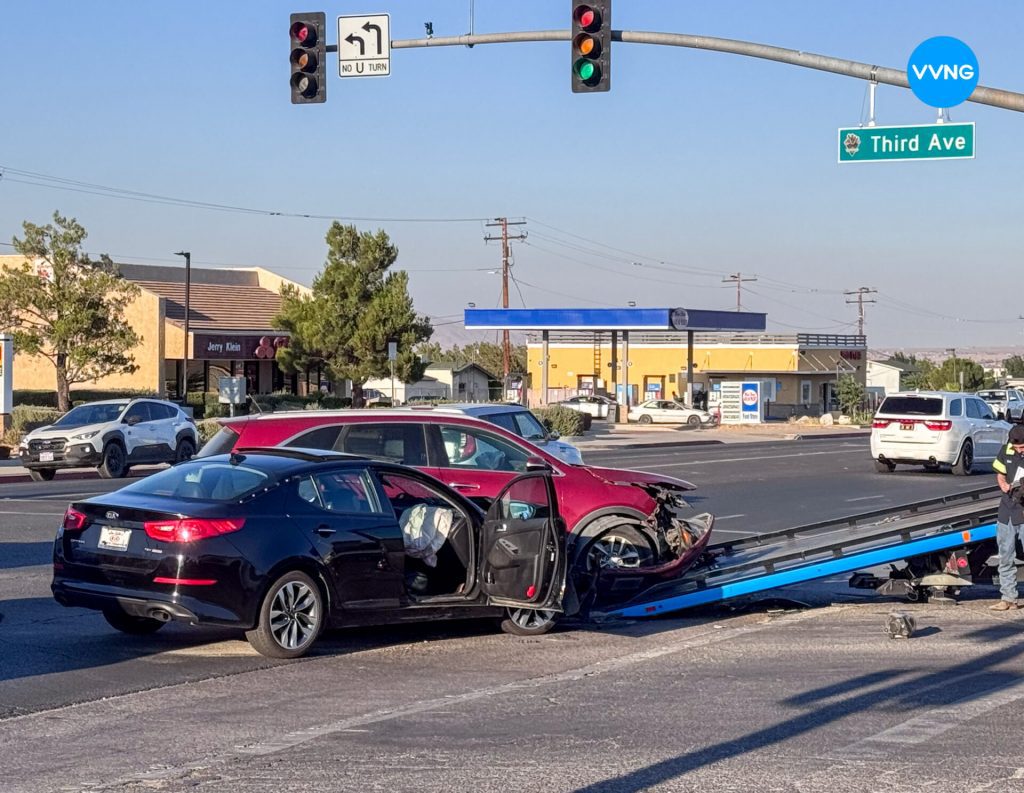 A busy intersection scene shows two damaged vehicles involved in a crash near a gas station and restaurant. A black sedan with deployed airbags and an open driver-side door is parked beside a maroon SUV with severe front-end damage, partially loaded onto a flatbed tow truck. A worker in a reflective vest stands nearby. Overhead traffic lights display red signals with a "No U Turn" sign and a green street sign reading "Third Ave." The sky is clear, and several other vehicles and businesses are visible in the background.