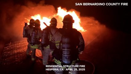 A team of San Bernardino County Fire firefighters in full turnout gear, including oxygen tanks, battles an intense residential structure fire at night in Hesperia, California. The flames blaze brightly ahead of them, casting an orange glow across the smoke-filled scene. The name “SAUCERMAN” is visible on one firefighter’s gear. At the top right, white text reads “SAN BERNARDINO COUNTY FIRE.” Below, bold white text at the bottom reads: “RESIDENTIAL STRUCTURE FIRE, HESPERIA, CA – APRIL 25, 2025.”