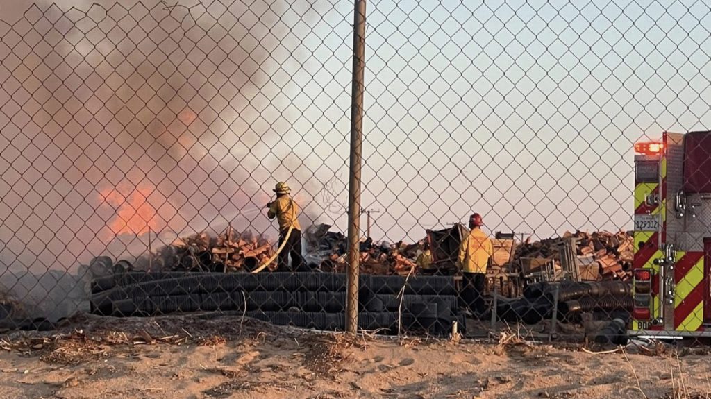 Two firefighters in yellow protective gear work to extinguish a fire burning through a large pile of wooden debris behind a chain-link fence. One firefighter is actively spraying water on the flames, while the other stands nearby. Thick smoke and bright orange flames rise from the burning pile. A fire truck with flashing lights is parked to the right, and stacks of pipes or hoses are visible in the foreground. The scene takes place on dry, sandy ground under a clear sky.