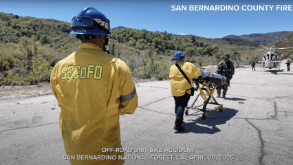 Firefighters and rescue personnel from San Bernardino County Fire assist with an off-road dirt bike accident. Two firefighters in yellow jackets, one with "SBCoFD" on the back, escort a patient on a stretcher toward a waiting medical helicopter on a cracked asphalt clearing. Armed rescue personnel stand nearby under a clear blue sky, surrounded by forested hills. Bold white text at the bottom reads: “OFF-ROAD DIRT BIKE ACCIDENT, SAN BERNARDINO NATIONAL FOREST, CA – APRIL 28, 2025.”