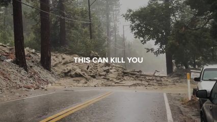 A rain-soaked mountain road is blocked by a large mudslide consisting of rocks, debris, and fallen trees. Power lines hang low over the scene, and visibility is reduced due to heavy rain and mist. Parked vehicles are visible on the right side of the road. Overlaying the image is bold white text that reads, ‘THIS CAN KILL YOU,’ warning of the dangers of mudslides and hazardous road conditions.