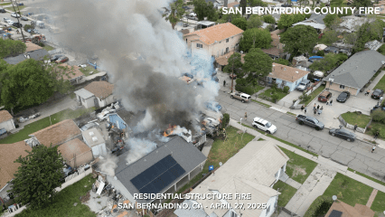 Aerial view of a residential neighborhood in San Bernardino, California, where a house is fully engulfed in flames with thick black smoke billowing into the sky. Firefighters are actively working on the scene, and multiple fire engines and emergency vehicles are parked on the surrounding streets. Nearby homes and residents are visible, with some people watching from their yards and driveways. The image includes the text: “RESIDENTIAL STRUCTURE FIRE – SAN BERNARDINO, CA – APRIL 27, 2025” and “SAN BERNARDINO COUNTY FIRE” in the top right corner.